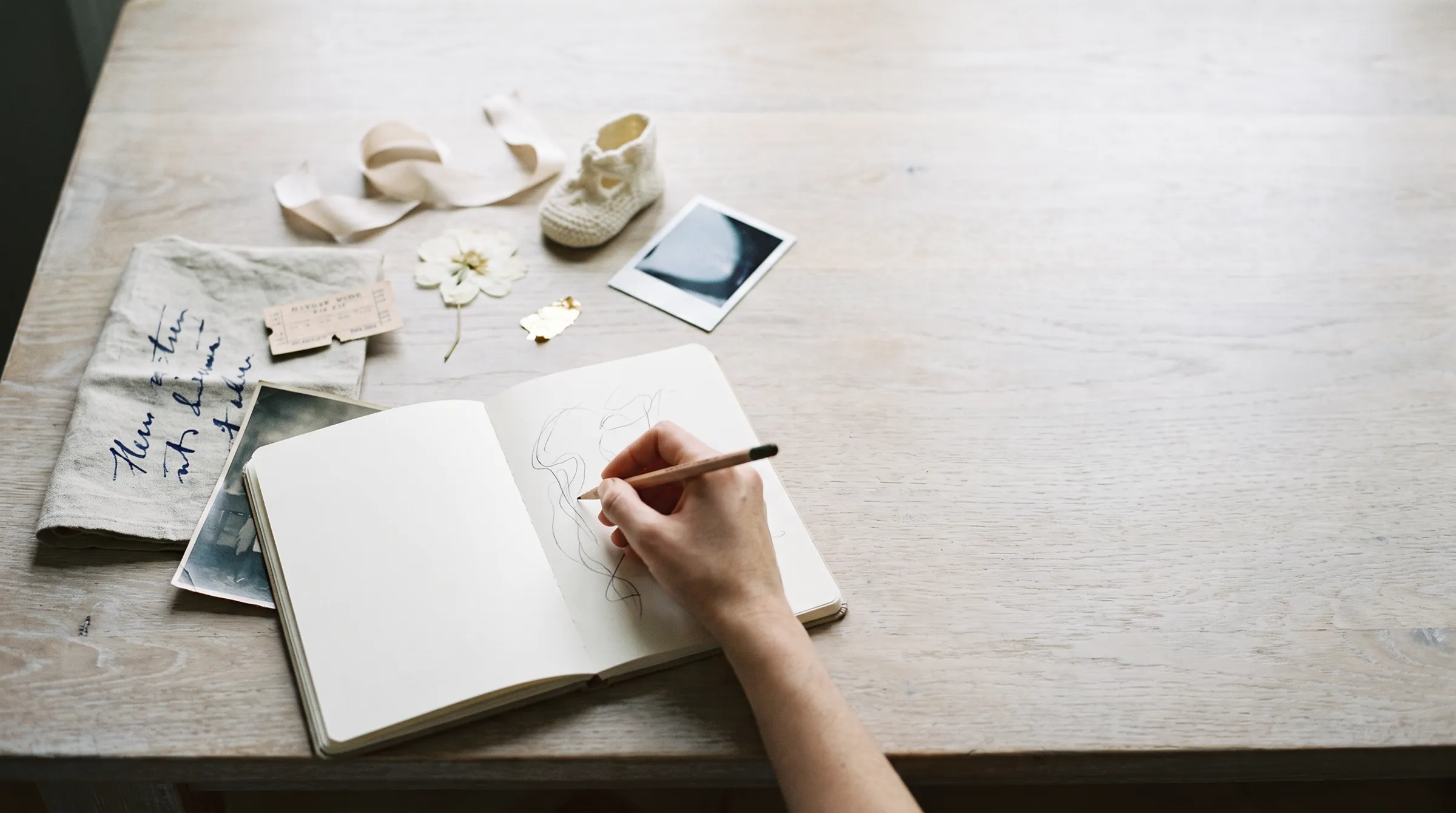Mesa del taller con recuerdos de una familia: un boleto, una flor seca, un zapatito tejido, una foto y una carta, y la mano de la artista dibujando en su cuaderno.